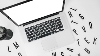 Nahaufnahme eines offenen weißen Laptops auf einem weißen Schreibtisch aus Vogelperspektive, umgeben von verstreuten schwarzen Buchstaben, die das Wort „WORDS“ bilden, neben einem schwarzen Stifthalter – in Schwarz-Weiß.