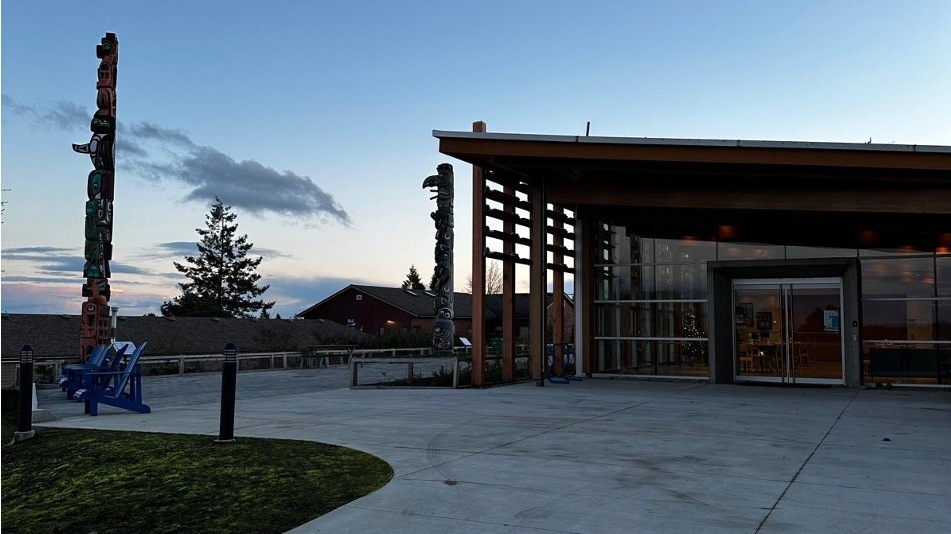 Moderner Gebäudeeingang mit Glasfassade und Holzelementen auf einem offenen Platz in der Dämmerung; im Vordergrund eine gepflasterte Fläche, links mehrere geschnitzte Totempfähle und blaue Sitzgelegenheiten, darüber ein klarer Abendhimmel.