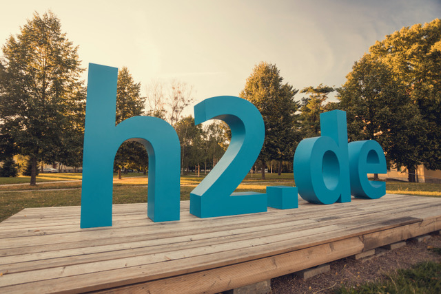 Große blaue 3D-Buchstaben h2.de auf einer Holzplattform im Park mit Bäumen im Hintergrund