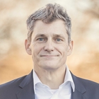 Ein Mann vor einem hellen Hintergrund. Er trägt ein weißes Hemd und ein schwarzes Anzugsoberteil.