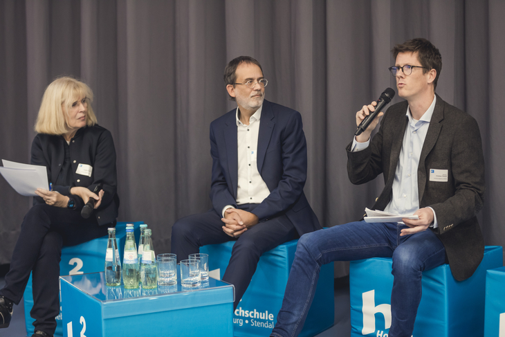 Drei Personen sitzen in einem Hörsaal und diskutieren vor Publikum.