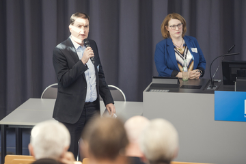 speaker is addressing an audience in a lecture theatre. Another speaker can be seen in the background.