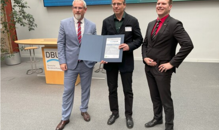 Three men are standing in the foreground. In the center is the working group member Ingolf Seick, holding a folder in his hands in a presentational manner, which contains the Innovation Award. The two other men stand beside him and are also smiling.