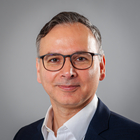 Ein Mann mit einer Brille vor einem grauen Hintergrund. Er trägt ein weißes Hemd und ein schwarzes Anzugsoberteil.