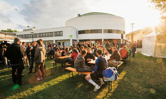 Vor dem Audimax der Hochschule sind Bänke, Tische und eine Bühne aufgebaut. Studierende, Lehrende und Gäste lauschen dem Orchester, lachen und unterhalten sich.