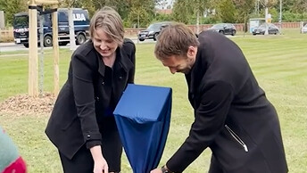 [Translate to English:] Die beiden Promovierten enthüllen das kleine Schild, das vor dem Baum steht.