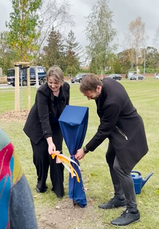 Die beiden Promovierten enthüllen das kleine Schild, das vor dem Baum steht.
