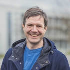Ein Mann lächelt. Er trägt ein hellblaues T-Shirt und eine dunkelblaue Jacke. Der Hintergrund ist verschwommen.