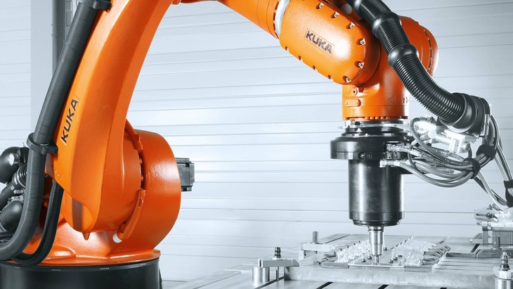 Orangefarbener Industrieroboter von KUKA, der mit einem Fräskopf über einem eingespannten Metallwerkstück arbeitet; gut erkennbar sind Roboterarm, Werkzeugaufnahme und der Bearbeitungstisch in einer industriellen Umgebung.