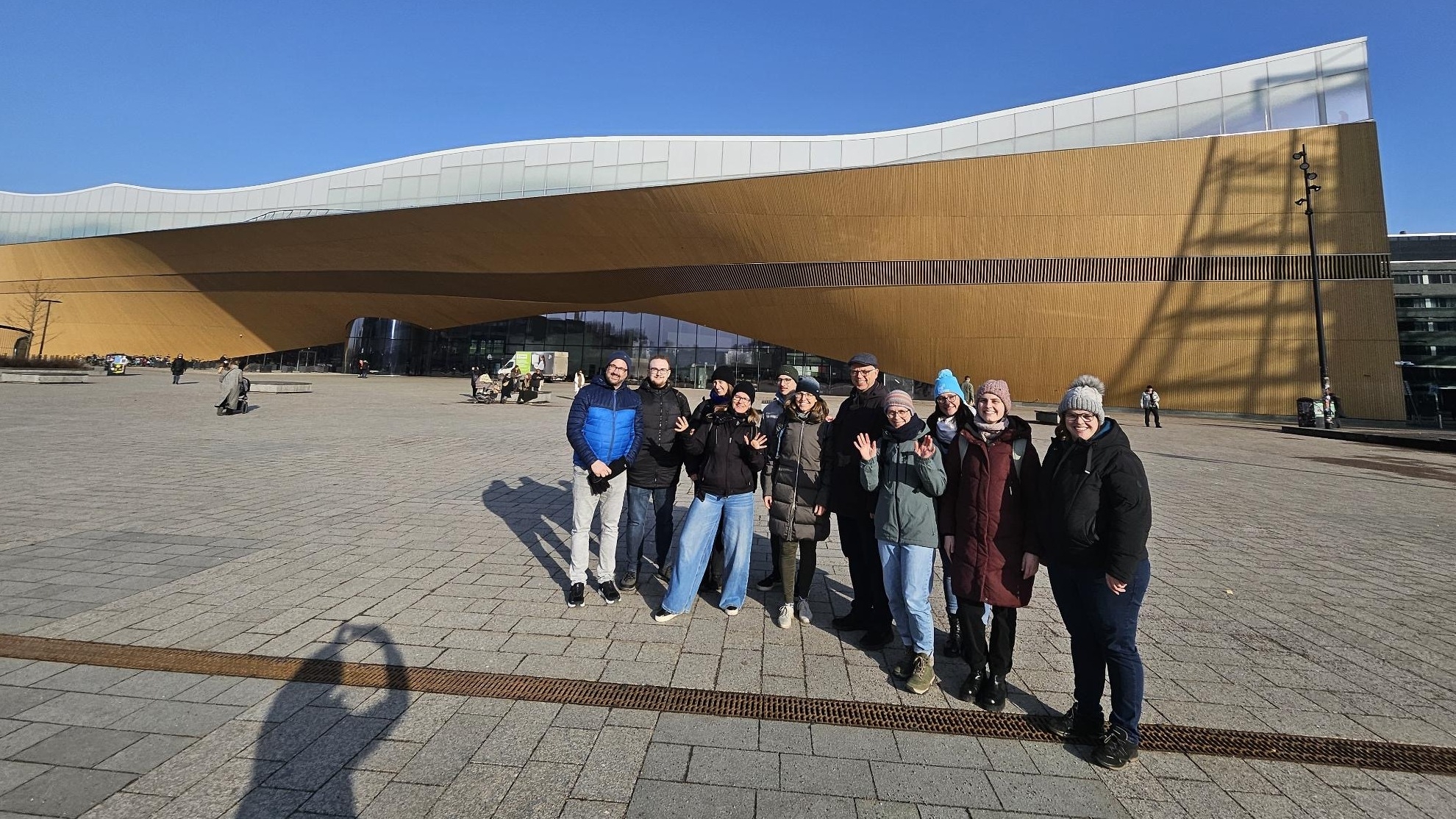 Eine Gruppe von Menschen steht auf einem großen Platz vor einem modernen goldfarbenen Gebäude und posiert für ein Foto.