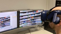 Ein Mann mit VR-Brille sitzt vor zwei Monitoren, auf denen Regale mit Produkten dargestellt sind, und erlebt eine virtuelle Supermarkt-Umgebung zur KI-gestützten Prognose von Kaufverhalten.