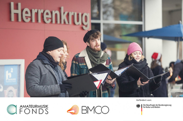 Foto mit vier singenden Personen vor der Herrenkrug Mensa, mit Logos der Förderer