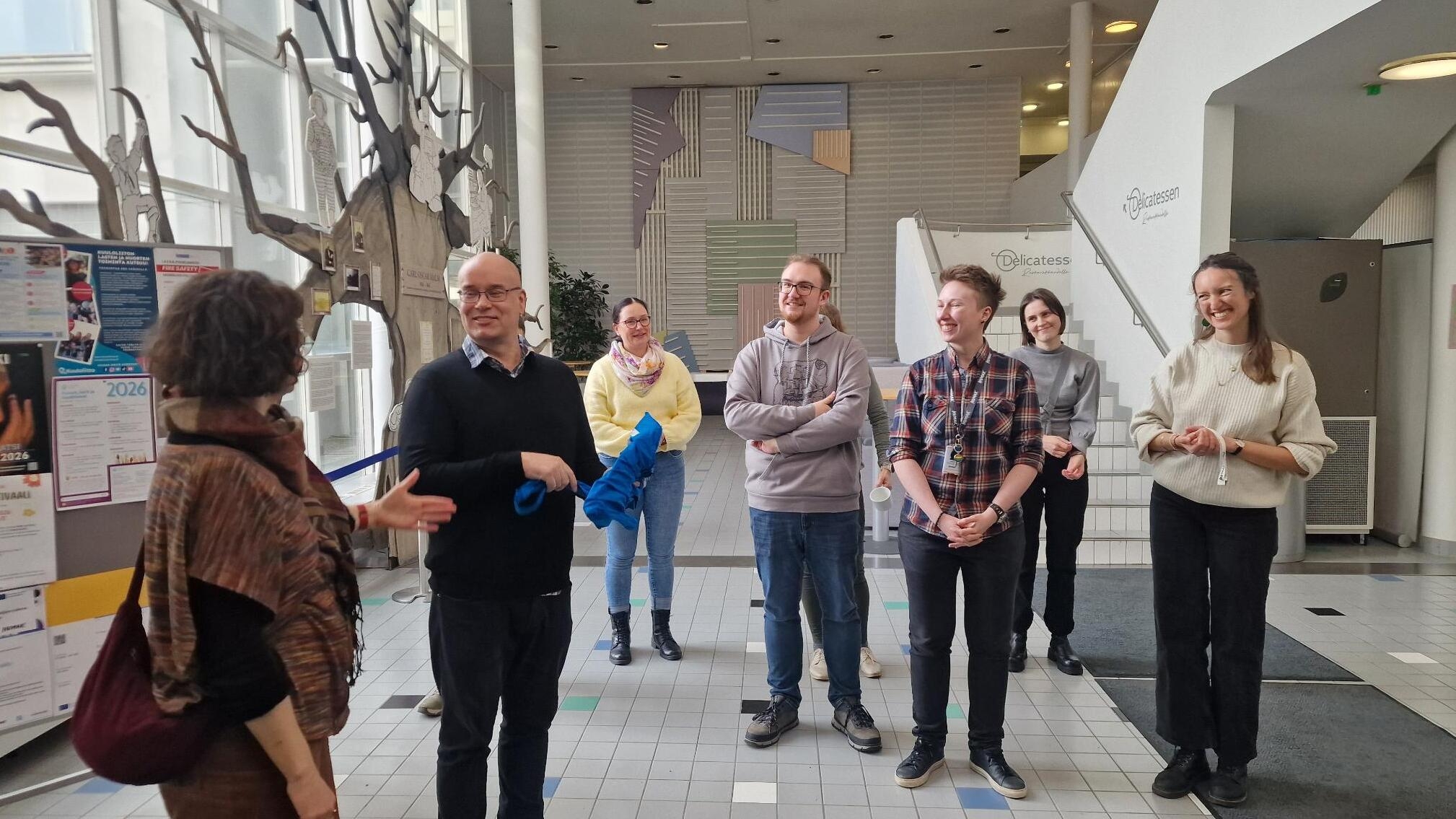Eine Person erklärt einer kleinen Gruppe in einem hellen Foyer etwas; die Teilnehmenden stehen im Halbkreis und hören zu.