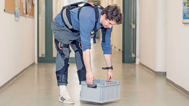 Eine Person demonstriert in einem Flur ein rückenunterstützendes Exoskelett und hebt eine Transportkiste an.