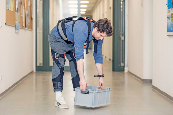 Eine Person demonstriert in einem Flur ein rückenunterstützendes Exoskelett und hebt eine Transportkiste an.
