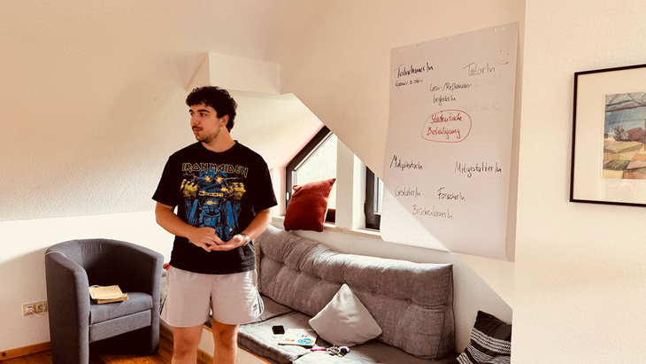 Person steht in einem Wohnraum neben einem Sofa; an der Wand hängt ein Flipchart mit Stichworten zu „Studentische Beteiligung“.