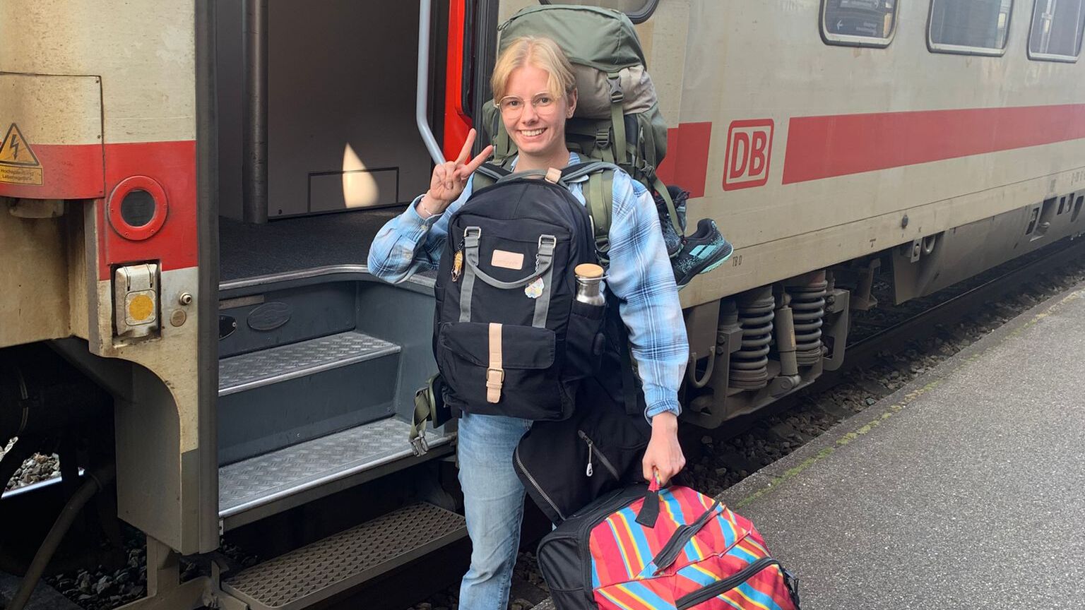 Person mit großem Rucksack und weiterem Gepäck steht auf einem Bahnsteig vor einem offenen Einstieg eines DB-Fernzugs, zeigt ein Victory-Zeichen und wirkt abfahrbereit für eine Reise.
