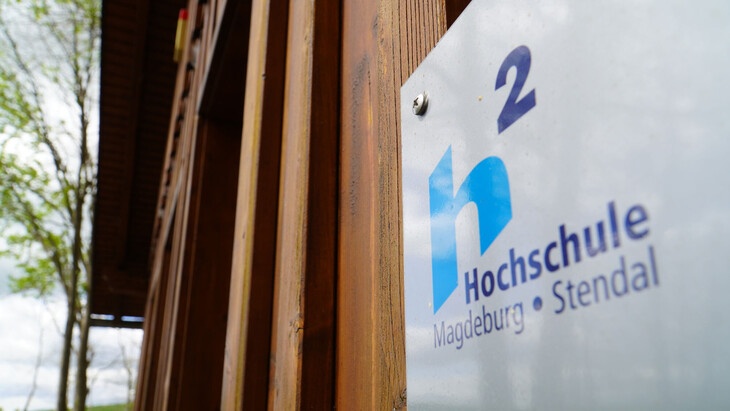 Schild mit Logo und Schriftzug „Hochschule Magdeburg · Stendal“ an einer Holzfassade, im Hintergrund Bäume und Grünfläche.