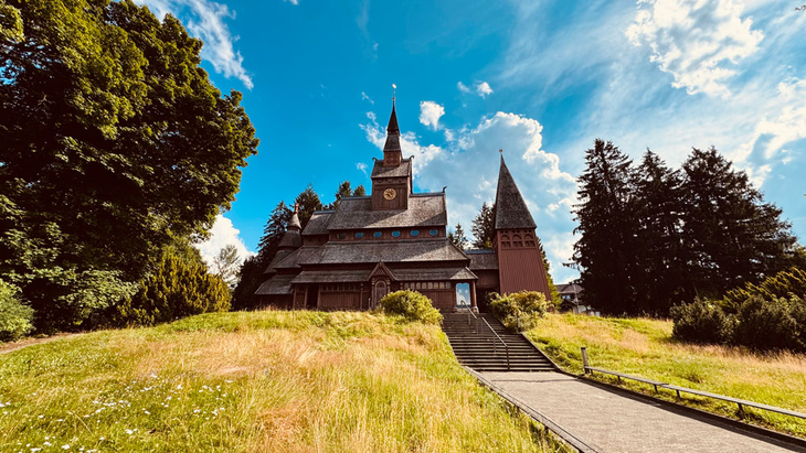 Holzkirche mit Uhrturm auf einer Anhöhe, umgeben von Bäumen und Wiesen unter blauem Himmel.
