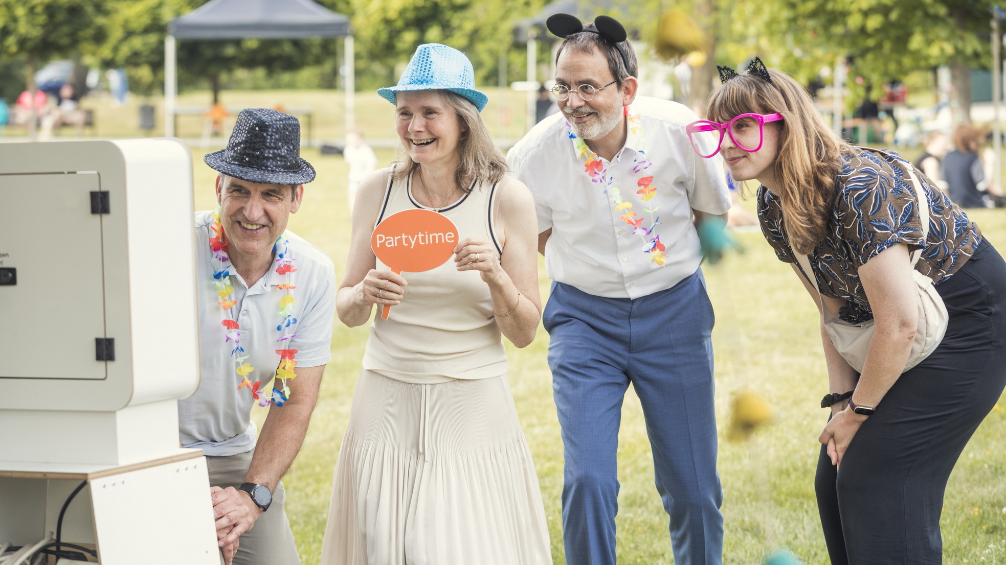 Vier verkleidete Personen posieren bei einem Outdoor-Fest fröhlich vor einer Fotobox, tragen bunte Hüte, Accessoires und Blumenketten, während im Hintergrund weitere Menschen auf der Wiese sitzen.