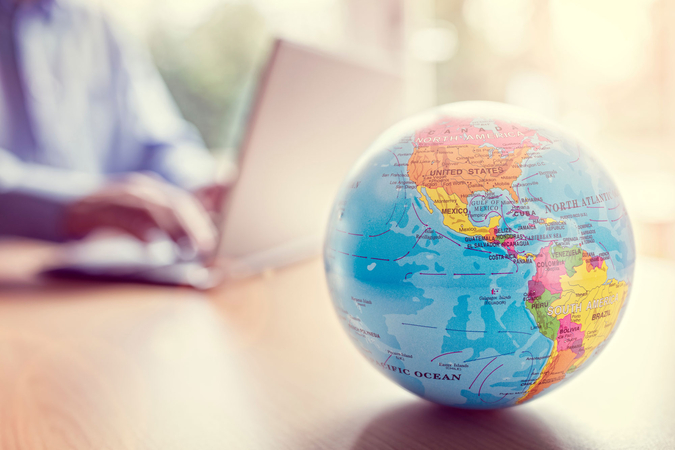 A colourful globe stands in the foreground on a desk, showing mainly North and South America. In the blurred background, a person is working on a laptop, suggesting the planning or organisation of international activities.