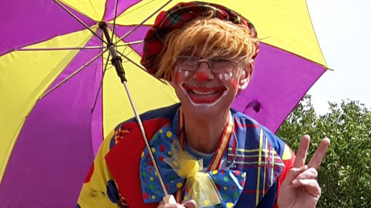 Eine als Clown verkleidete Person hält einen bunten Regenschirm und zeigt ein Peace-Zeichen.