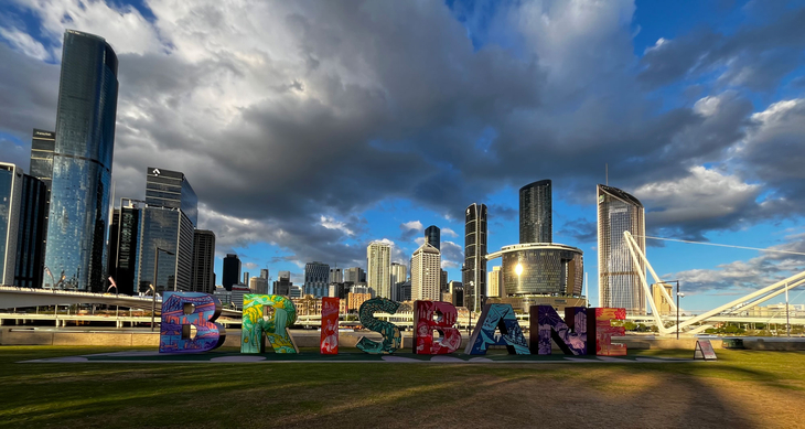 Panorama of the Brisbane skyline with modern skyscrapers, dramatic cloudy skies and colourful, large ‘BRISBANE’ letters in the foreground on a green space.