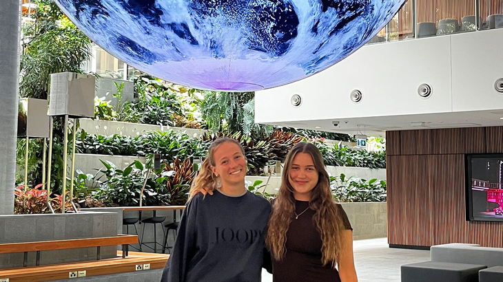 Two young women stand smiling in a modern, light-filled foyer beneath a large, illuminated globe installation, surrounded by lots of green plants and seating.
