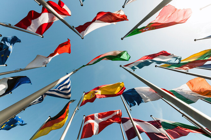 Several colourful national flags flutter in the wind on tall flagpoles, photographed from a worm's-eye view against a clear blue sky. The different colours and patterns of the flags symbolise international diversity and cooperation.