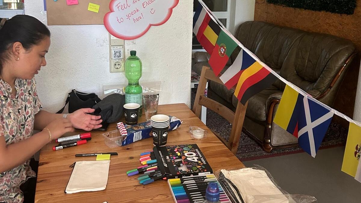 Ein Aufenthaltsraum mit einem großen Holztisch, auf dem Stifte, Haftnotizen und Bastelmaterial liegen. An der Wand hängt ein Plakat mit der Aufschrift „Write HERE your wishes for the next semester“ und bunten Haftnotizen. Daneben sind eine Girlande mit internationalen Flaggen, ein Sofa und eine sitzende Person am Tisch zu sehen.
