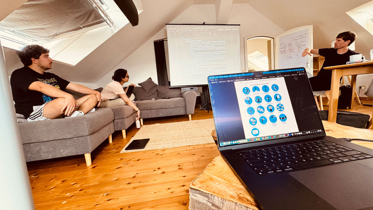 Strategietreffen in einem Wohnzimmer mit mehreren Personen auf Sofas und am Tisch, Präsentationsleinwand im Hintergrund und Laptop im Vordergrund.