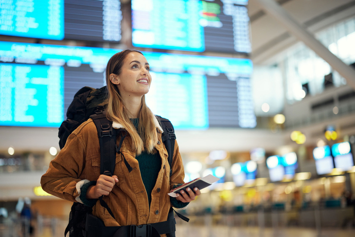 A young traveller with a large rucksack stands in an airport hall and looks up at the blue departure boards in the background. She holds her travel documents in her hand, while the brightly lit check-in counters and monitors blur around her.