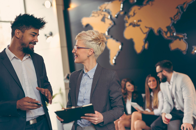 Several people in business attire are conversing in a modern office or conference area. In the foreground, a man and a woman are talking, both holding tablets or documents. In the blurred background, other people are seated on chairs, while a large stylised world map hangs on the wall, suggesting an international context and global cooperation.