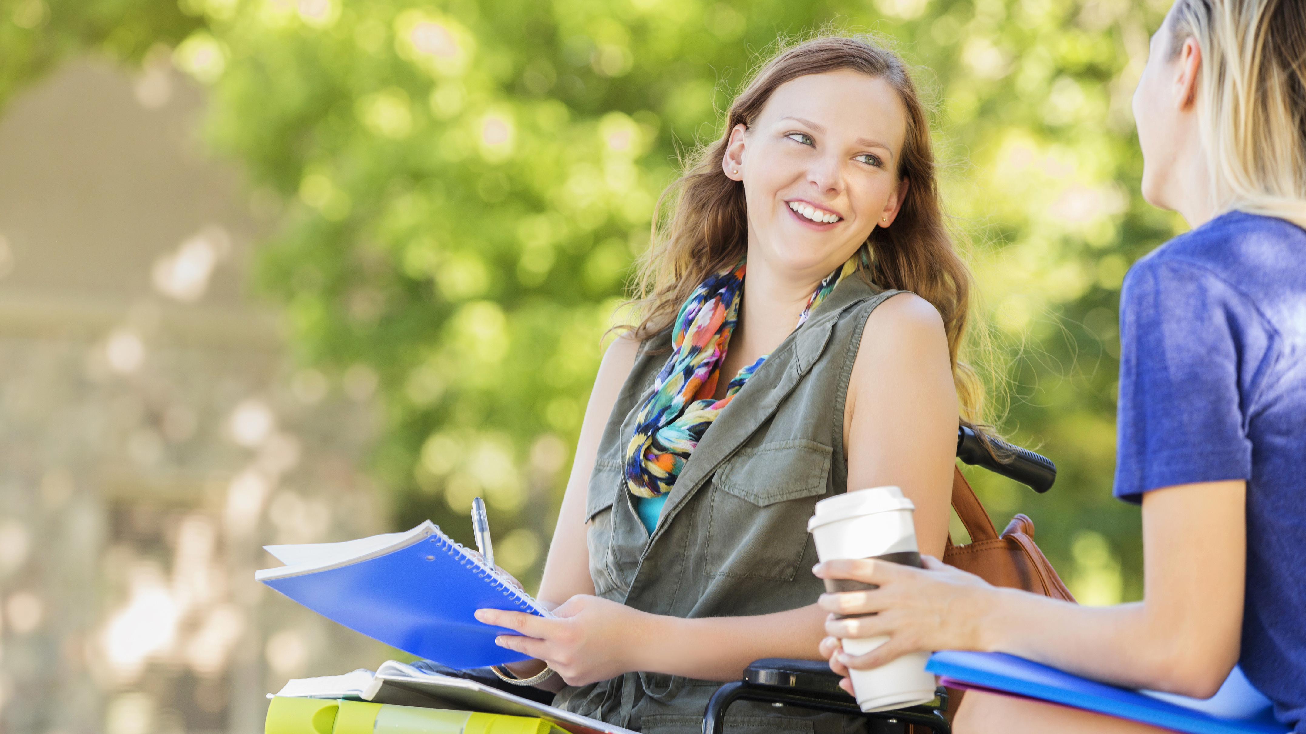 Junges Mädchen im Rollstuhl sitzt im Freien und unterhält sich mit einer anderen Person; sie hält Unterlagen auf dem Schoß, wirkt entspannt und ist von grüner, unscharfer Parklandschaft umgeben.