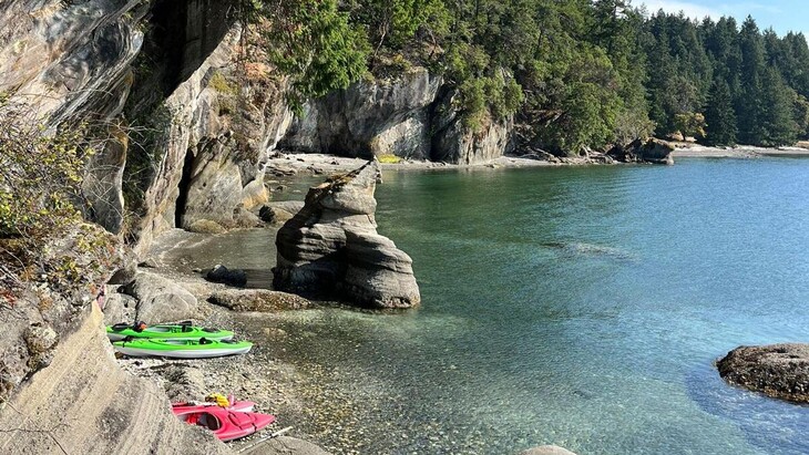 Felsige Küstenbucht mit klarem türkisblauem Wasser, steilen bewaldeten Klippen und mehreren bunten Kajaks, die am schmalen Kiesstrand unter überhängenden Felsen liegen.