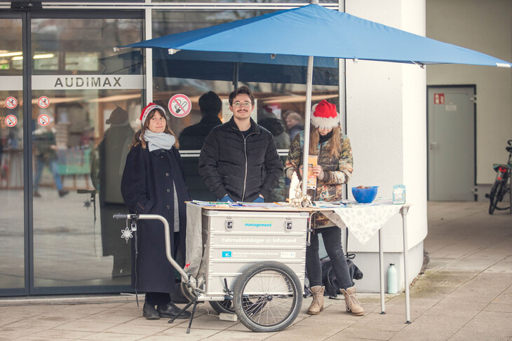 Drei Personen stehen vor einem Gebäude an einem Infostand unter einem blauen Sonnenschirm; auf dem Tisch liegen Flyer und Materialien.