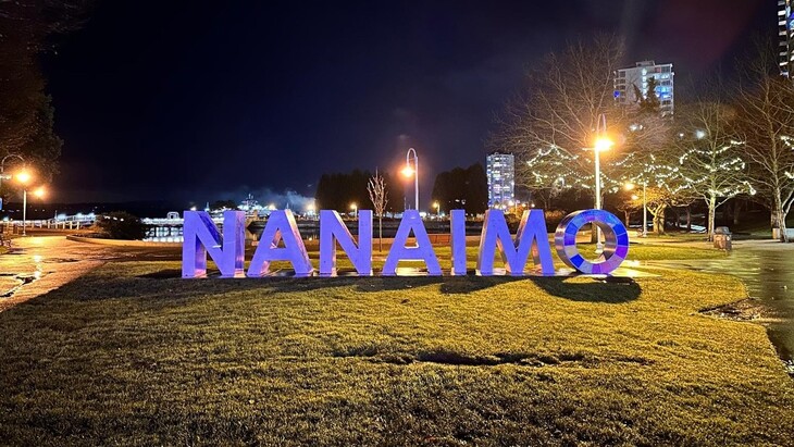 Nachtaufnahme eines Parks mit großen, violett beleuchteten Buchstaben „NANAIMO“ im Vordergrund, umgeben von Laternen und Bäumen mit Lichterketten, im Hintergrund mehrstöckige Wohngebäude vor dunklem Himmel.