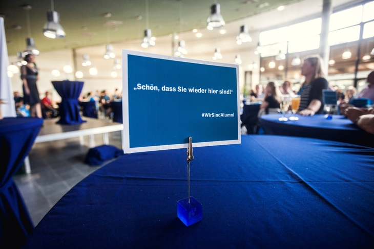 Auf einem Tisch steht eine blaue Postkarte mit dem Schriftzug "Schön, dass Sie wieder hier sind." und "Wir sind Alumni".