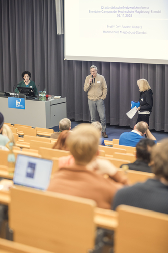 Ein Vortragender spricht in einem Hörsaal vor Publikum. Links ist eine weitere Vortragende zu sehen. Am rechten Bildrand steht die Moderatorin der Veranstaltung.