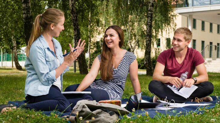 Zwei Studentinnen und ein Student sitzen vor einem Hochschulgebäude auf einer Decke im Rasen und diskutieren über ihren Studienunterlagen
