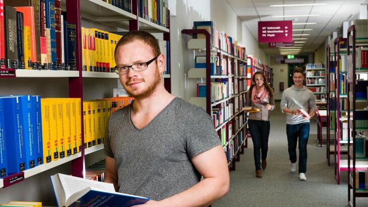Ein Student liest an ein Regal gelehnt ein Buch, im Hintergrund laufen zwei Studierende den Gang entlang