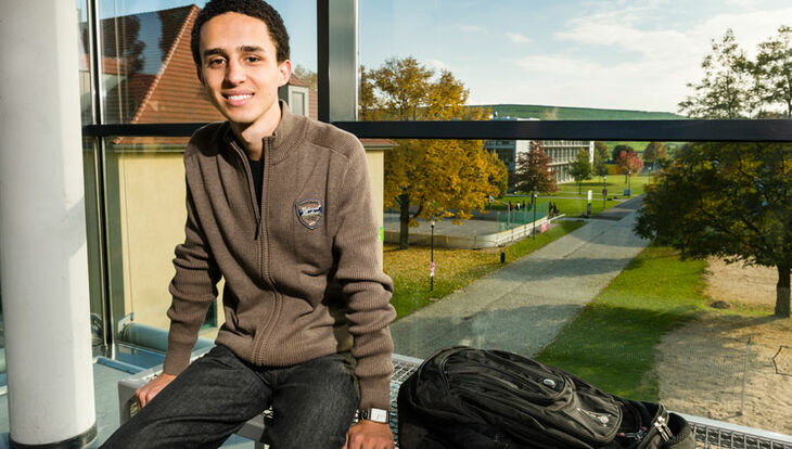 Ein brasilianischer Austauschstudent sitzt im Glasbalkon des International Office und lächelt freundlich in die Kamera, im Hintergrund sieht man den Campus mit Laborhallen und Beachvolleyballfeld