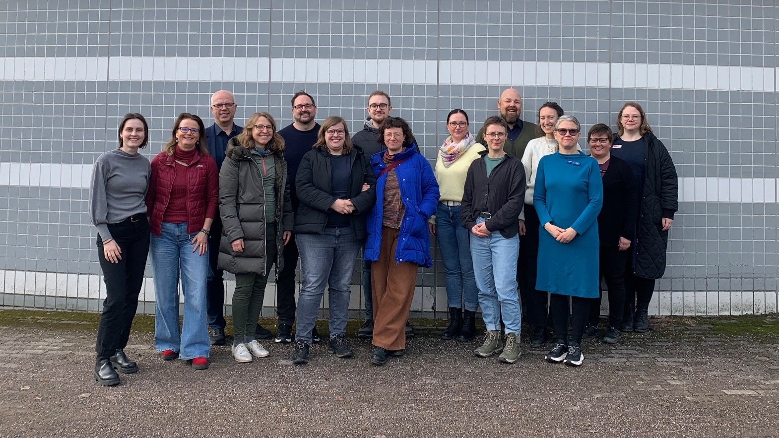 Eine Gruppe von Menschen steht vor einer grauen Gebäudefassade mit der Aufschrift „VALKEA TALO“ und posiert für ein Gruppenfoto.