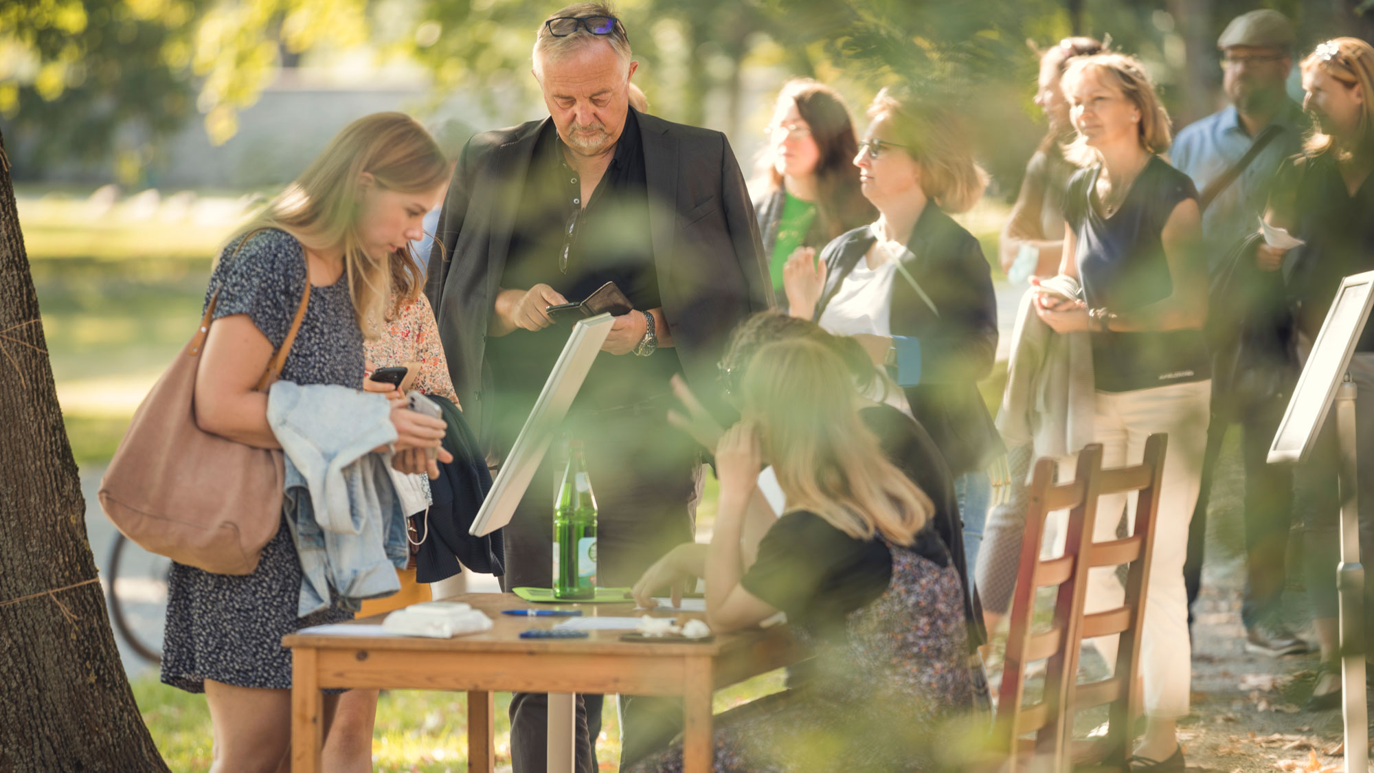 Menschen stehen bei einer Registrierung im Freien an einem Tisch mit Formularen und Stiften; im Hintergrund sind weitere Teilnehmende in einer parkähnlichen Umgebung zu sehen.