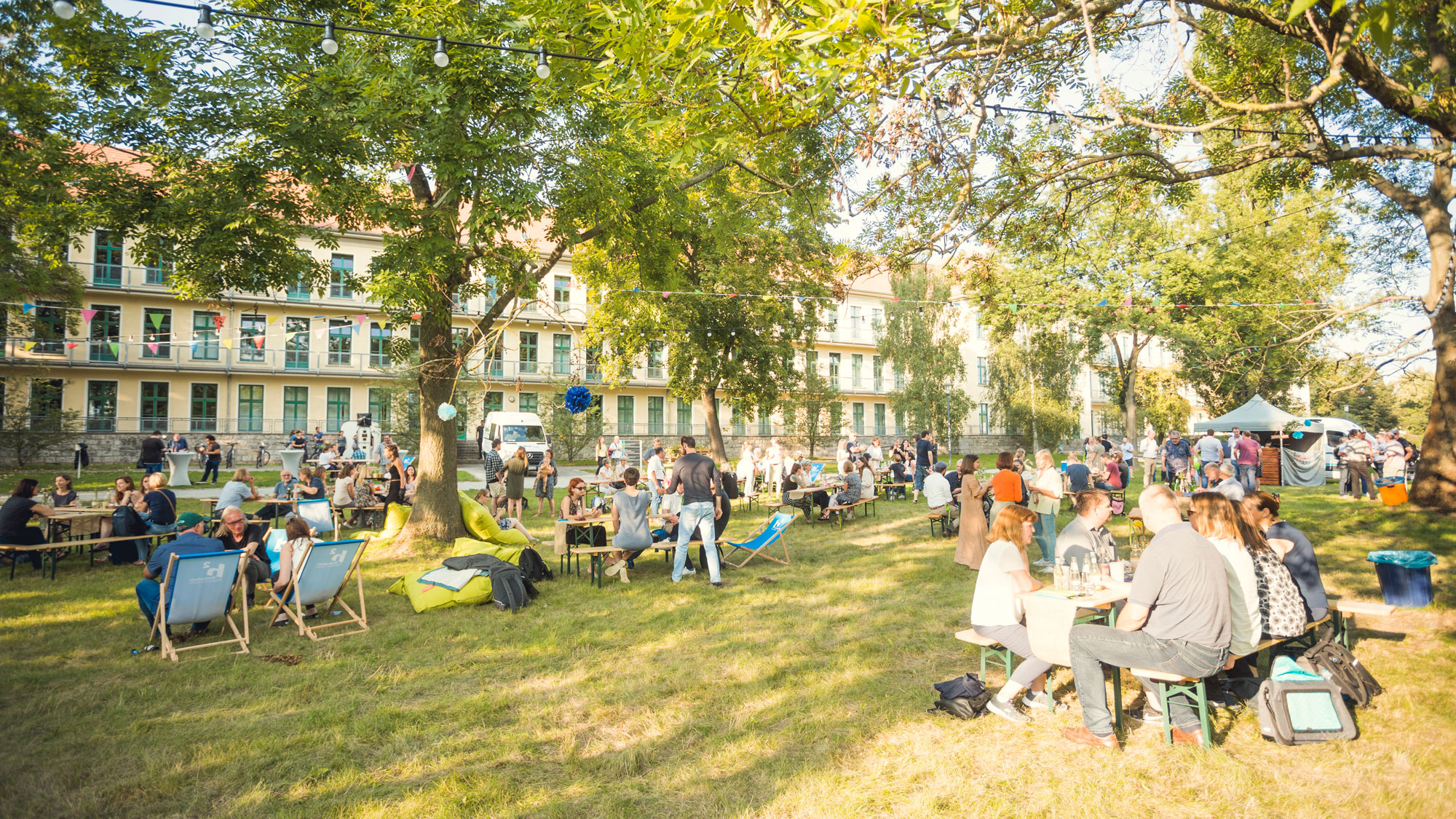 Weitläufige Garten- oder Parkszene mit vielen Menschen, die an Tischen und Liegestühlen auf einer Wiese unter Bäumen zusammenkommen; im Hintergrund steht ein Gebäude, darüber hängen Lichterketten.