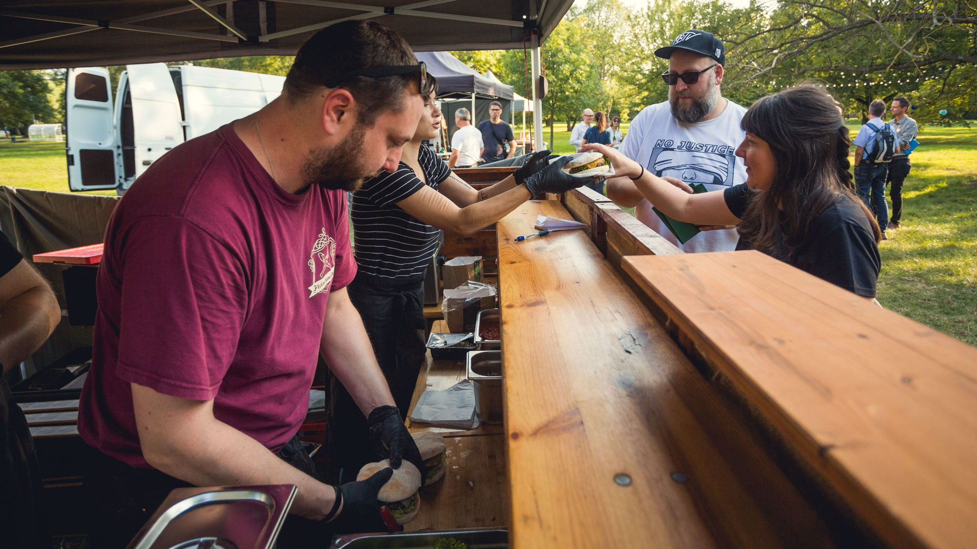 Mehrere Personen geben an einem Food-Stand im Freien Burger oder Sandwiches über eine Holztheke weiter; im Hintergrund stehen weitere Besucher im Park.