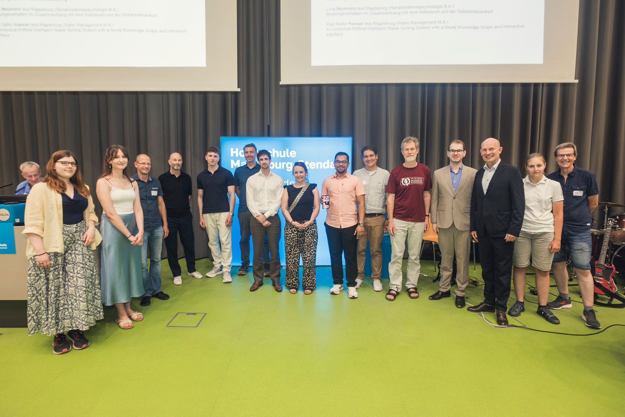 Eine größere Gruppe von Menschen steht auf einer Bühne vor Vorhängen und einem Hochschulbildschirm für ein gemeinsames Gruppenfoto. Im Hintergrund sind außerdem zwei Projektionsflächen sowie Musik- und Bühnenequipment zu sehen.