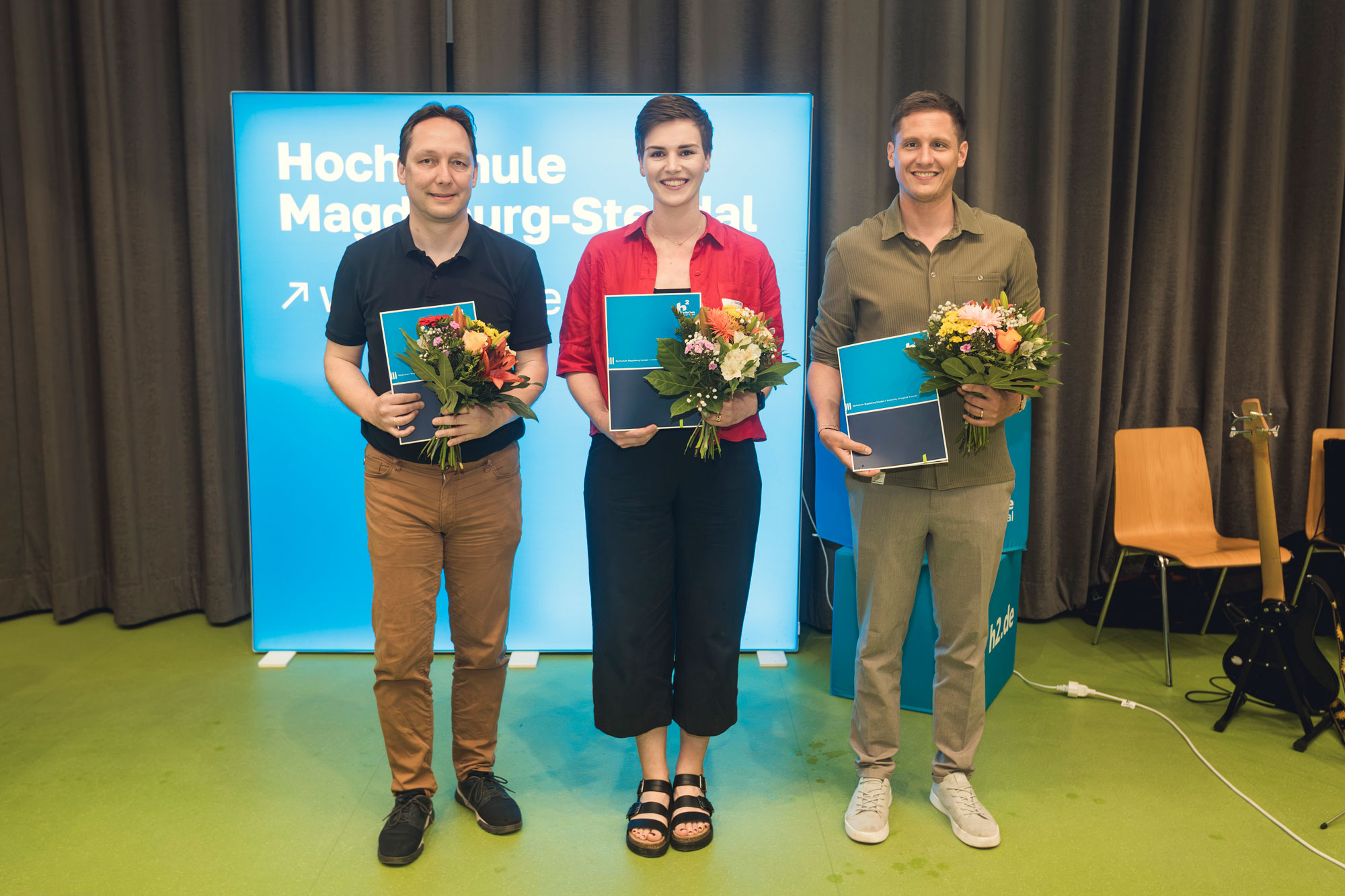 Drei Personen stehen bei einer Veranstaltung auf einer Bühne vor einem blauen Hochschul-Hintergrund, jeweils mit einem Blumenstrauß und einer Mappe in der Hand. Im Hintergrund sind Vorhänge, ein Stuhl und ein Instrument zu sehen.