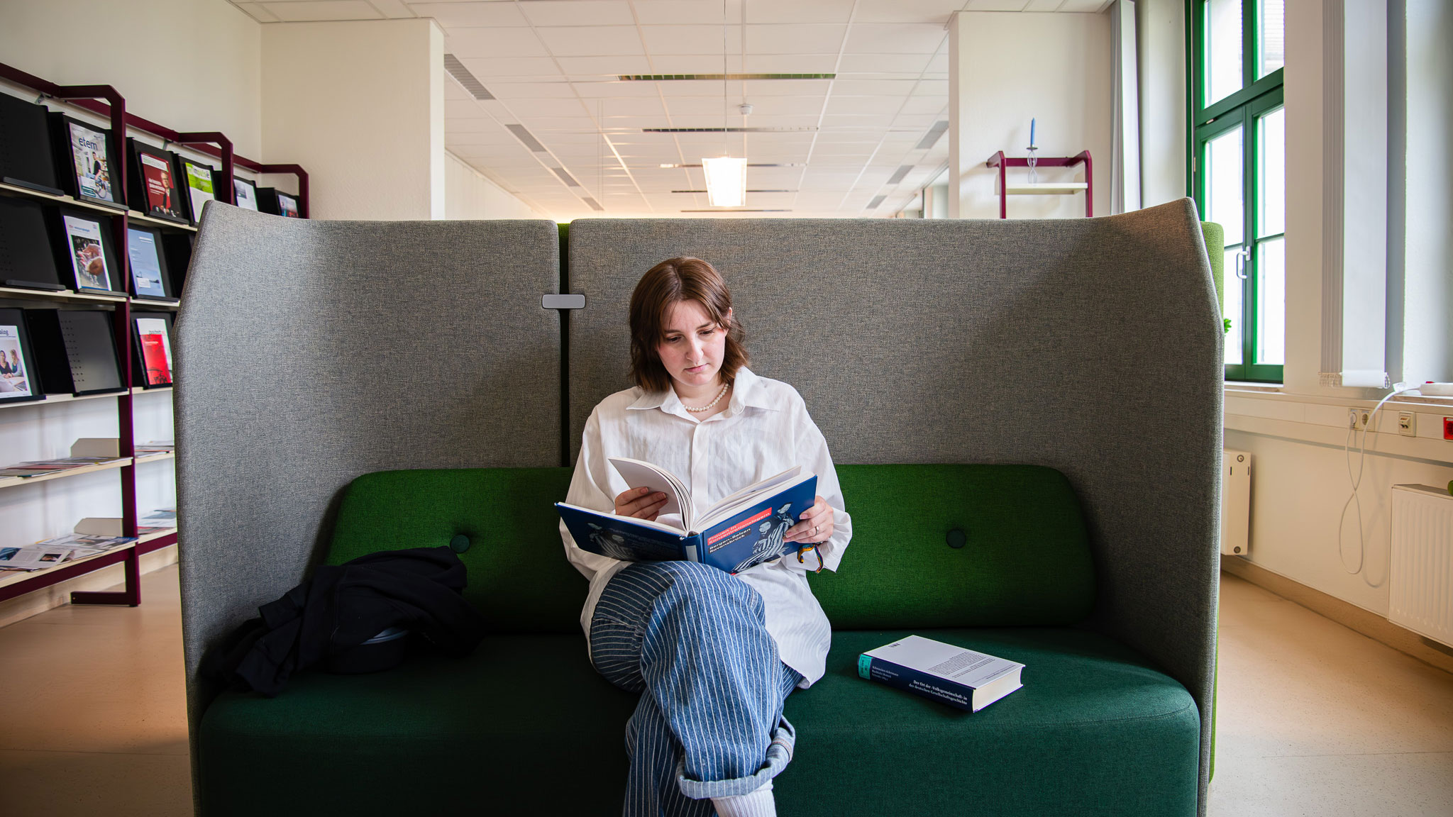 Eine Frau sitzt in einer Bibliothek auf einer grünen Sitzbank mit hohen, grauen Rückenlehnen und liest ein Buch. Neben ihr liegen ein weiteres Buch und eine Jacke; im Hintergrund sind Bücherregale und große Fenster zu sehen.