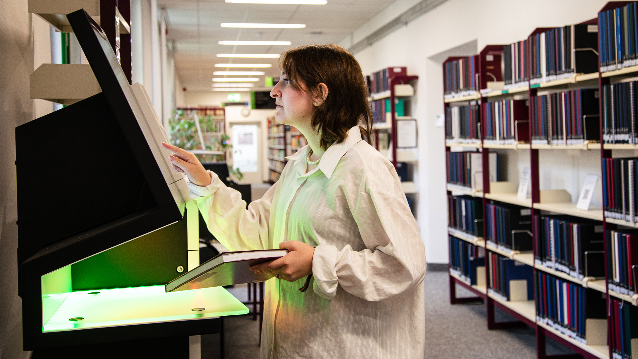 Eine Frau in einem Bibliotheksraum bedient ein Geräte-Terminal mit grün leuchtendem Licht und hält ein Buch in der Hand. Im Hintergrund stehen hohe Regale mit Büchern.